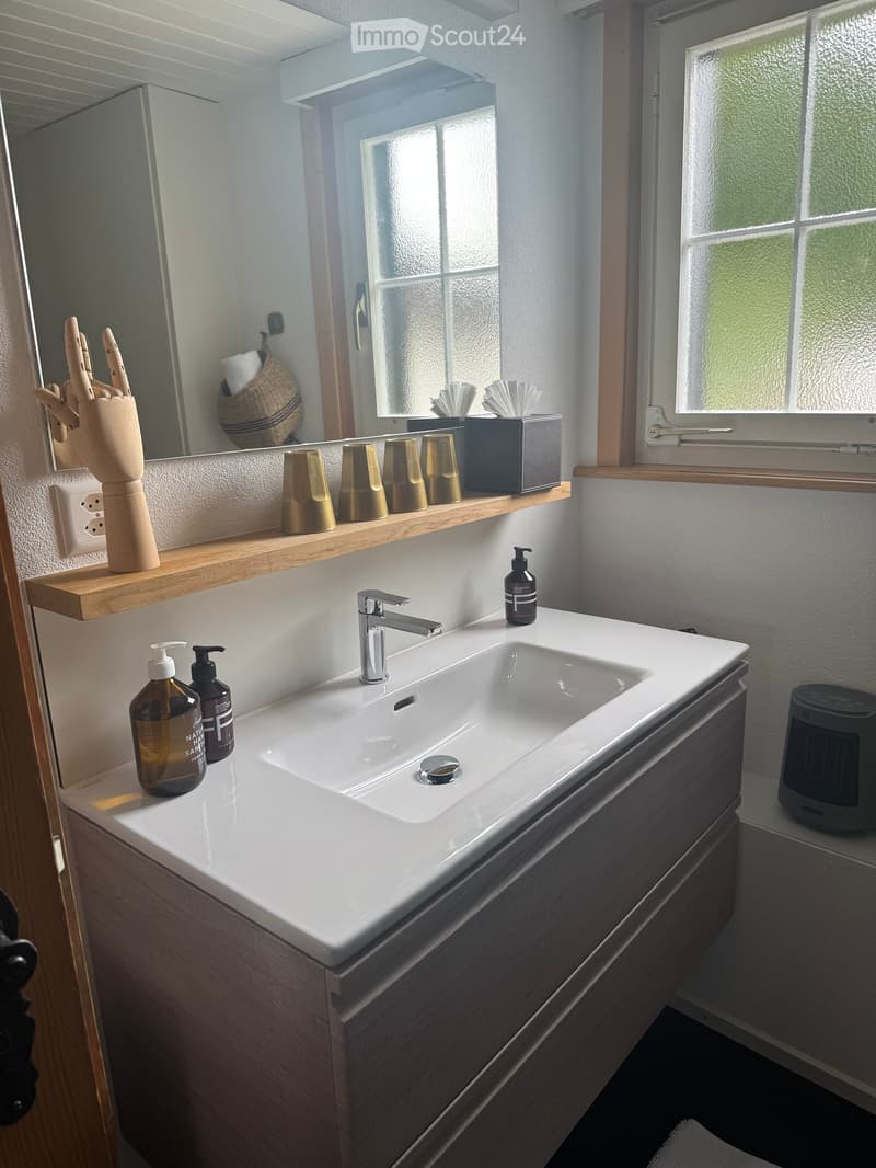 Modern bathroom with a white sink, chrome faucet, wooden shelf holding cups and a figurine, and a mirror above the sink. Bottles are placed on the counter.