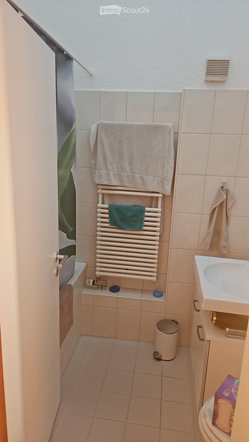 Compact bathroom with tiled walls and floor, featuring a sink, radiator with towels, toilet, and a small trash bin.