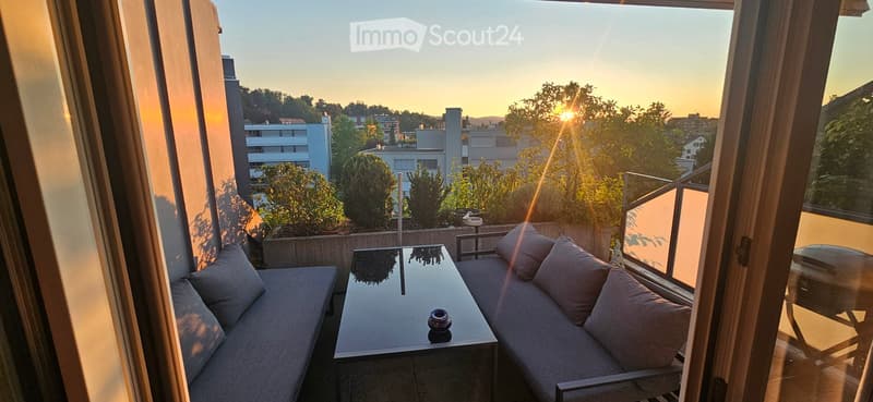 Balcony with a view of the city at sunset, featuring a grey sofa, a glass coffee table, and decorative pillows.