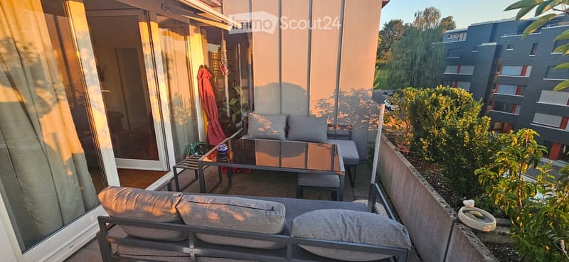 Balcony with a sofa, coffee table, and chairs, overlooking a modern building and greenery.
