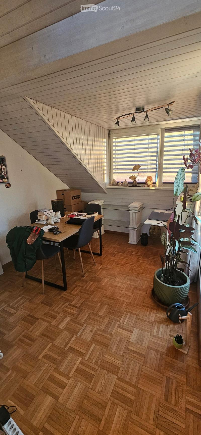 A cozy attic room with wooden flooring, slanted wooden ceiling, and large window with blinds. Contains a desk with a laptop, books, and a backpack, along with a large potted plant and decorative vases on the windowsill.