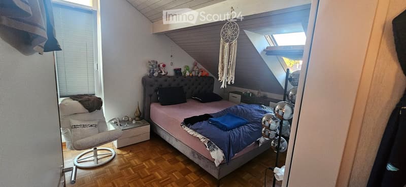 Cozy bedroom with a slanted ceiling, featuring a double bed with pink bedding, a plush chair, nightstand, and decorative items including a dreamcatcher and toys on the headboard.