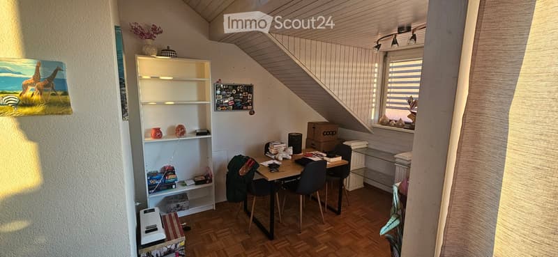 A cozy, slanted-ceiling room with a wooden floor, a small table with chairs, a white shelf with decorative items, and a painting of giraffes on the wall.