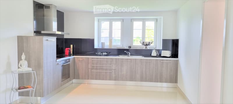 Modern kitchen with light wood cabinets, stainless steel appliances, and a sleek black backsplash. Features include a refrigerator, oven, sink, faucet, toaster, teapot, and decorative items on the counter and side table.