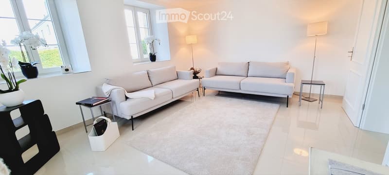 Modern living room with white walls, light-colored sofas, a white rug, and large windows letting in natural light. Includes a side table, floor lamp, and decorative vases.