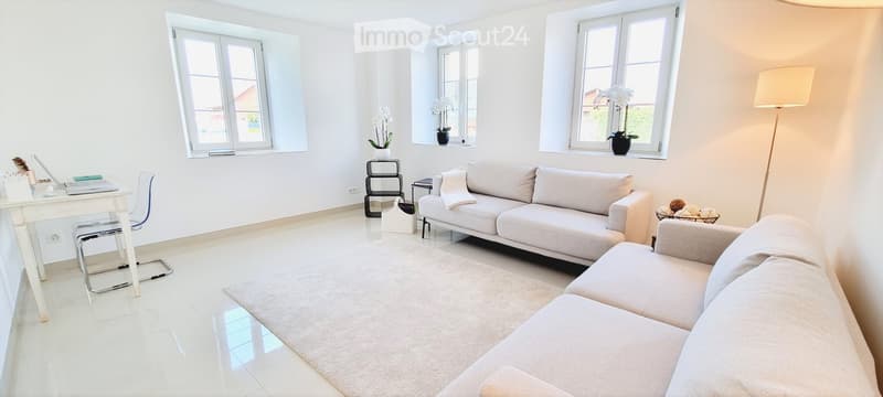 Modern living room with white walls, large windows, a beige sofa, a white chair, a small table with a laptop, a floor lamp, and decorative vases on the windowsills.