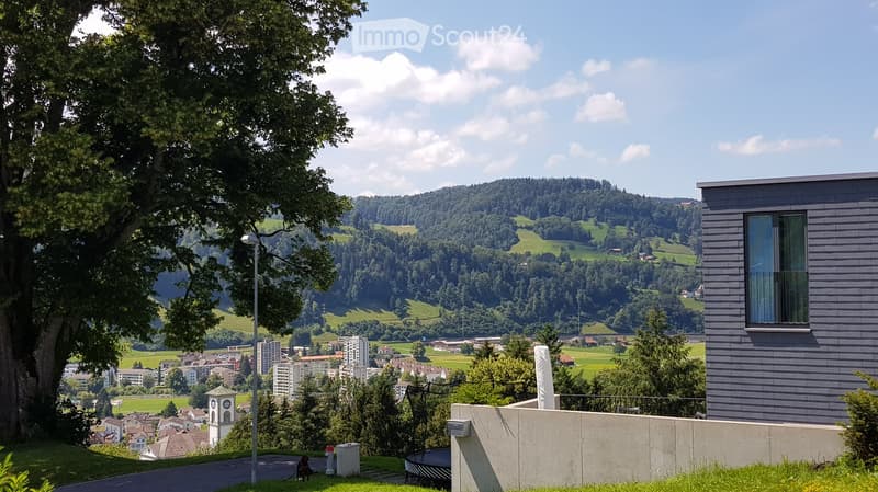 A scenic outdoor view shows a modern building with large windows and a balcony overlooking a lush green valley, hills, and a town with scattered houses and a church tower.
