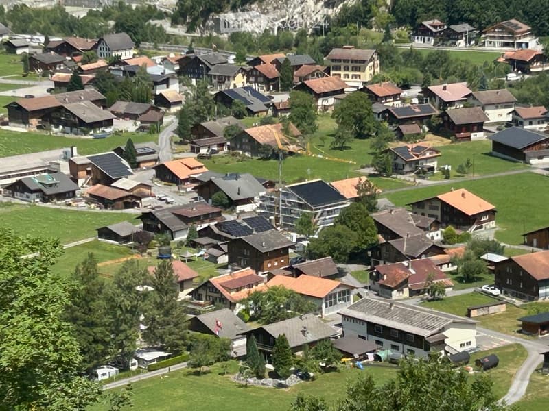 Aerial view of a small village with numerous houses, green lawns, and scattered trees. The houses have varied roof styles and colors, and the area is surrounded by mountains.