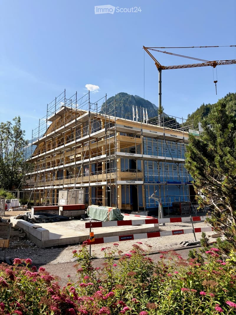 Multi-story building under construction with scaffolding, wooden framing, and visible signage. The site includes construction materials, a crane, and a mountainous backdrop.