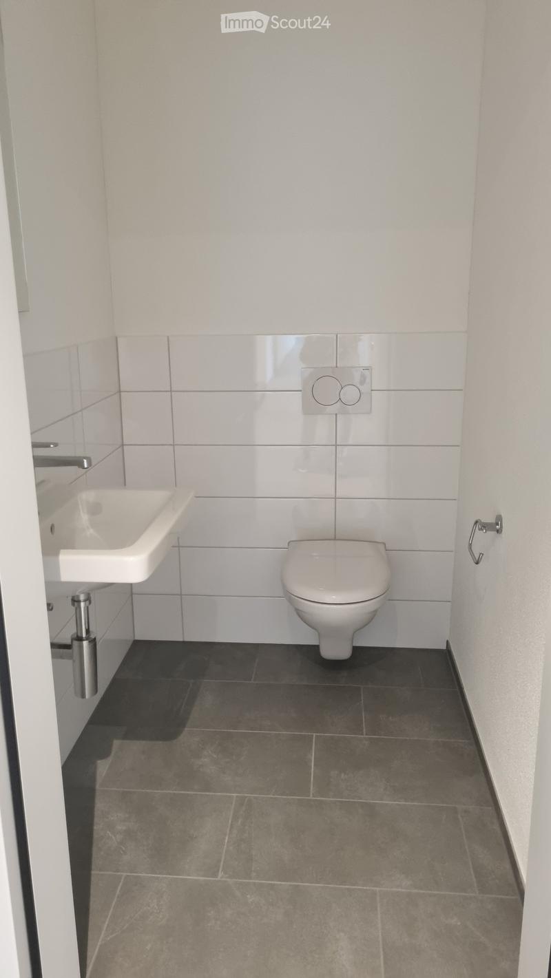 Modern bathroom with white tiled walls, a toilet, a sink, and dark grey tiled flooring.