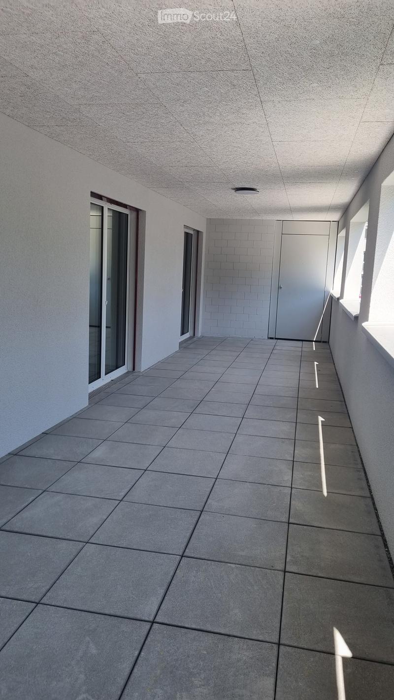 A clean, modern hallway with tiled flooring, white walls, and a ceiling light fixture. Features a glass door on the left and a closed door at the end.