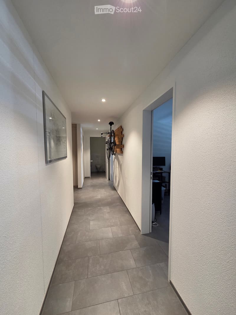 A narrow hallway with tiled flooring, white walls, and recessed lighting. A bicycle is mounted on the wall, and a mirror is visible on the left side. An open doorway leads to a room with a desk and computer.