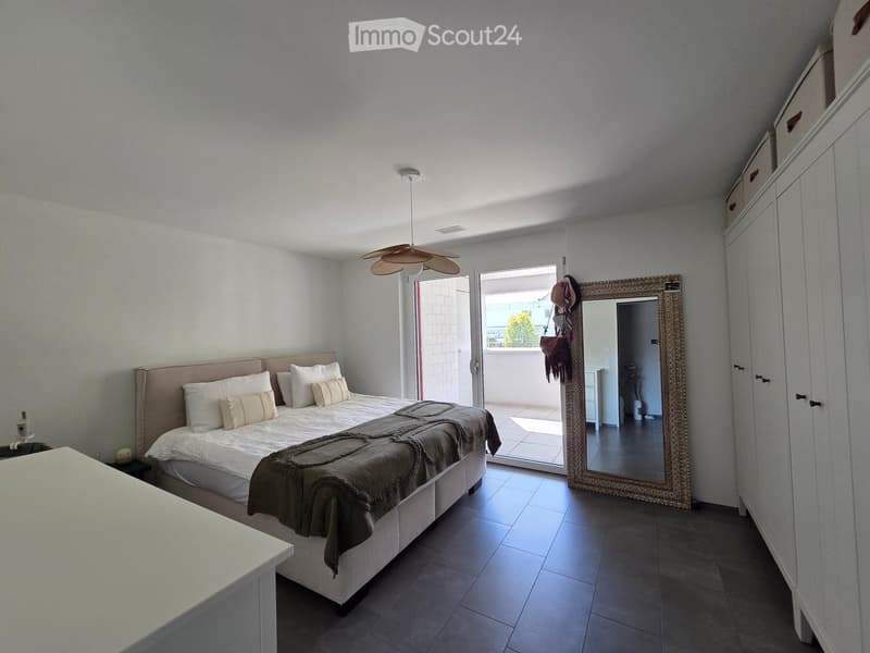 Modern bedroom with a large bed, nightstand, wardrobe, and a large mirror. The room features a fan hanging from the ceiling and an open door leading to a balcony.
