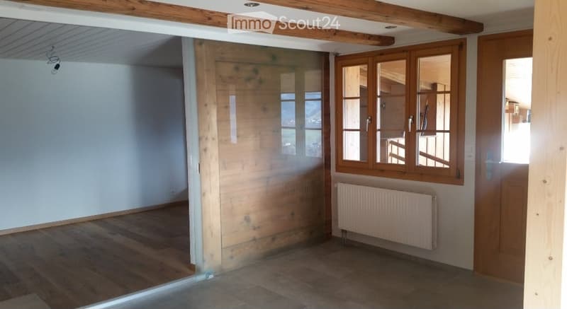 Empty room with wooden flooring, white walls, large window with wooden frame, radiator, and exposed wooden beams on the ceiling.