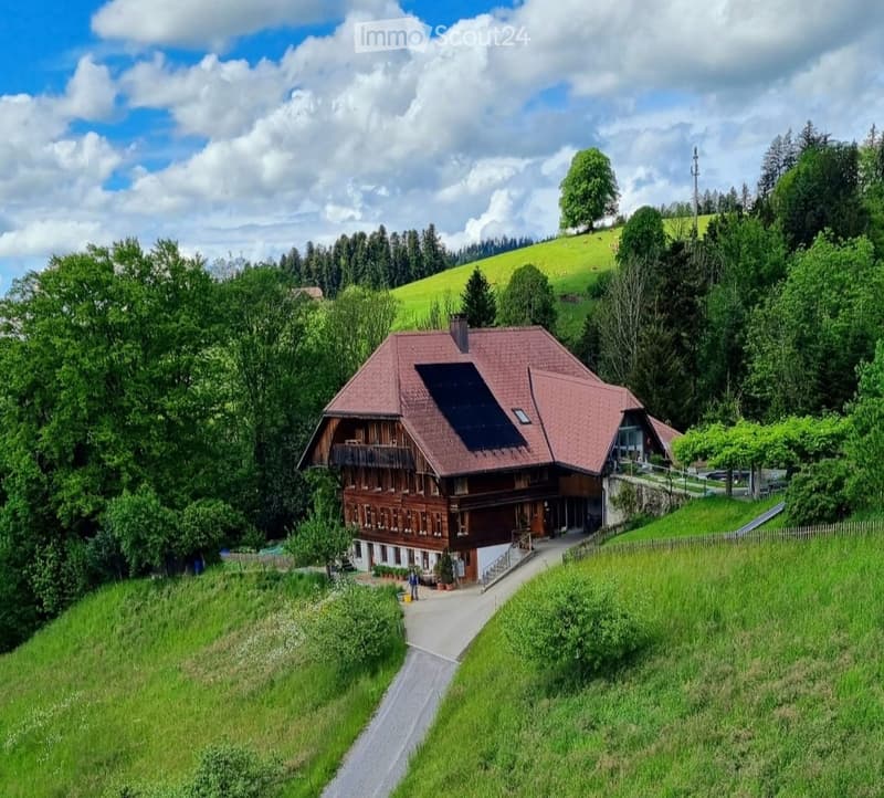 A two-story wooden house with a red-tiled roof, surrounded by lush greenery and hills.