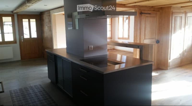 Modern kitchen with dark cabinets, a central island, and a built-in stove with a range hood. The space features wooden walls and large windows allowing natural light to fill the room.