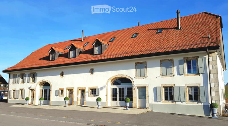 Two-story house with white walls, red-tiled roof, multiple windows with shutters, arched entrance, and potted plants.