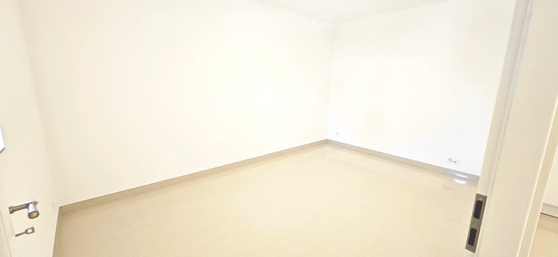 Empty room with white walls and light-colored flooring, featuring a partially open door and a visible door handle.