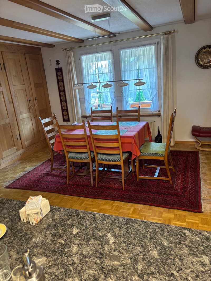 Dining area with wooden table, chairs, red table runner, and a large window with curtains. Wooden floor, decorative rug, and a small stool near the wall.