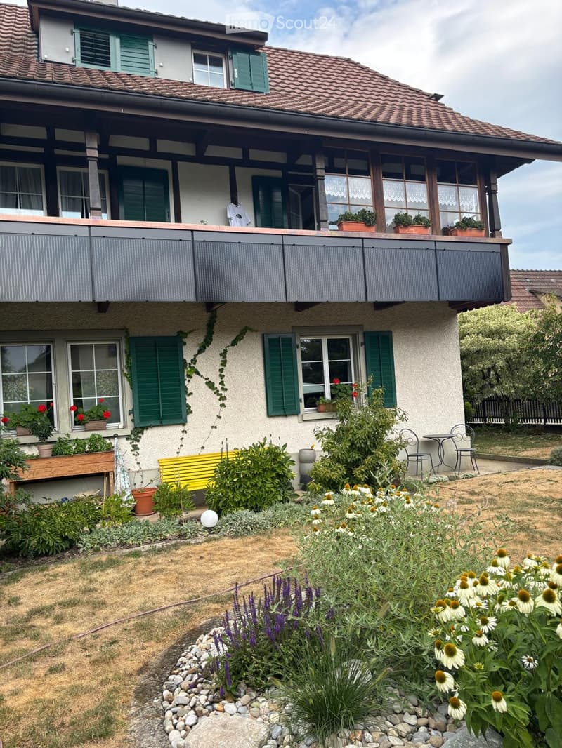 Two-story house with a tiled roof, green shutters, and a balcony with potted plants. The garden features a yellow bench, small table, chairs, and various flowers and shrubs.
