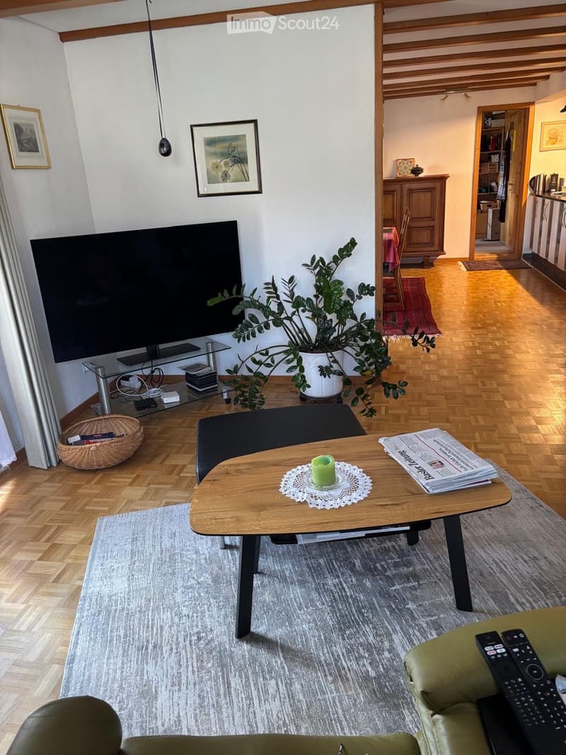 Living room with wooden flooring, grey sofa, coffee table, small table with a candle, plant, TV, and a view into the kitchen area.