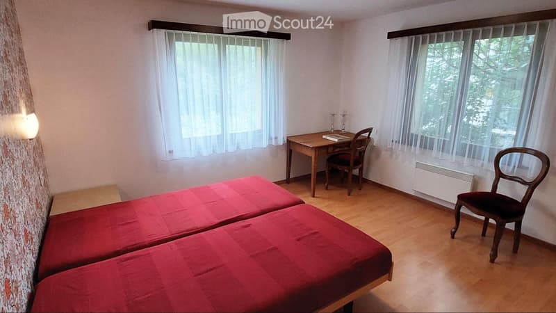 Cozy room with two single beds, a wooden desk, a chair, and a candle holder on the desk. Light-colored walls and wooden flooring.