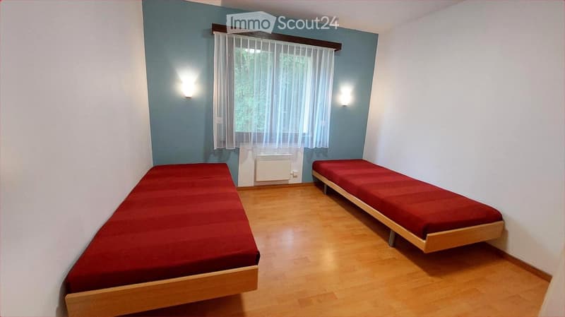 Two single beds with red bedding, wooden floor, white walls, window with sheer curtains, wall-mounted light fixtures, radiator.