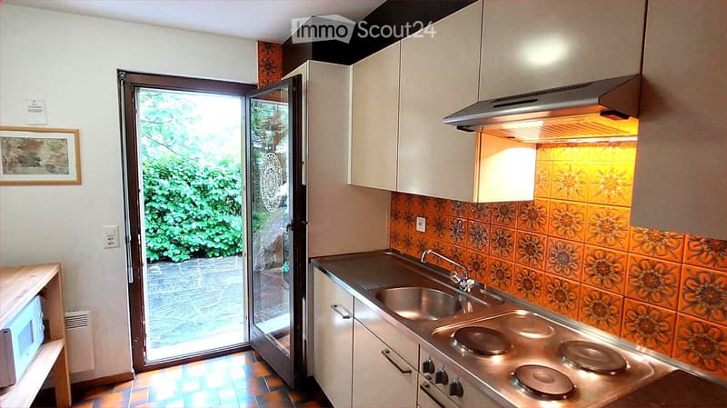 Modern kitchen with white cabinets, stainless steel sink and stove, and a microwave on a wooden counter. The backsplash features orange floral tiles, and a sliding glass door opens to a green outdoor area.
