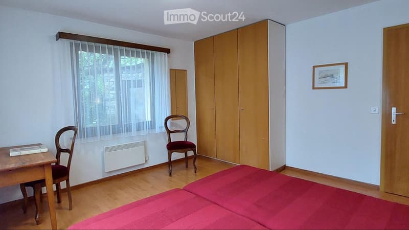 A small, furnished bedroom with a red bedspread, wooden floor, a wooden table with a book, two chairs, a built-in wardrobe, and a radiator under a window with sheer curtains.