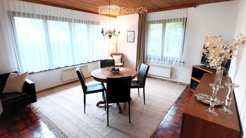 Bright living room with wooden ceiling, large windows, sofa, dining table with chairs, TV stand, and decorative vase with flowers.