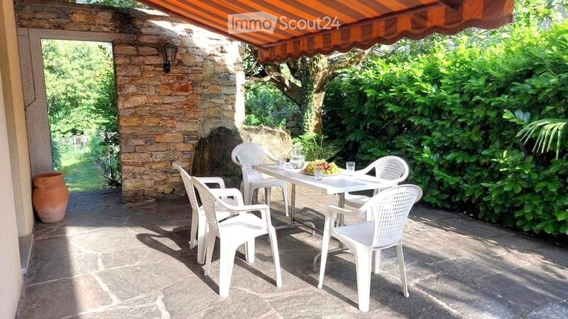 Outdoor patio with stone walls, white plastic chairs, and a table set with glasses and a bowl of fruit, shaded by an orange awning.