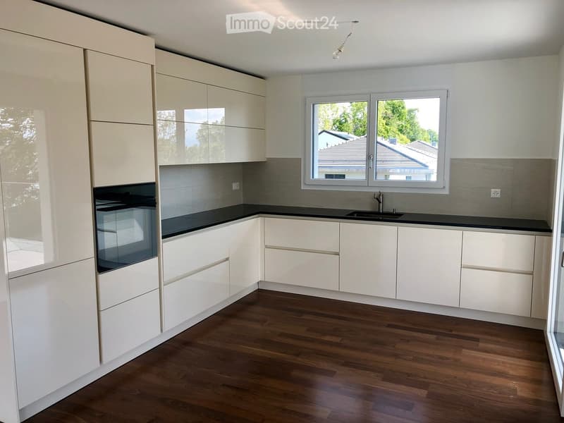 Modern kitchen with white cabinets, black countertops, and a wooden floor. Features a sink with a modern faucet and large windows overlooking a residential area.