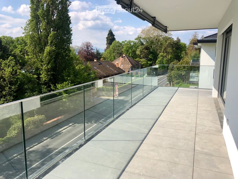 Modern balcony with glass railing, concrete floor, and scenic view of trees and houses.