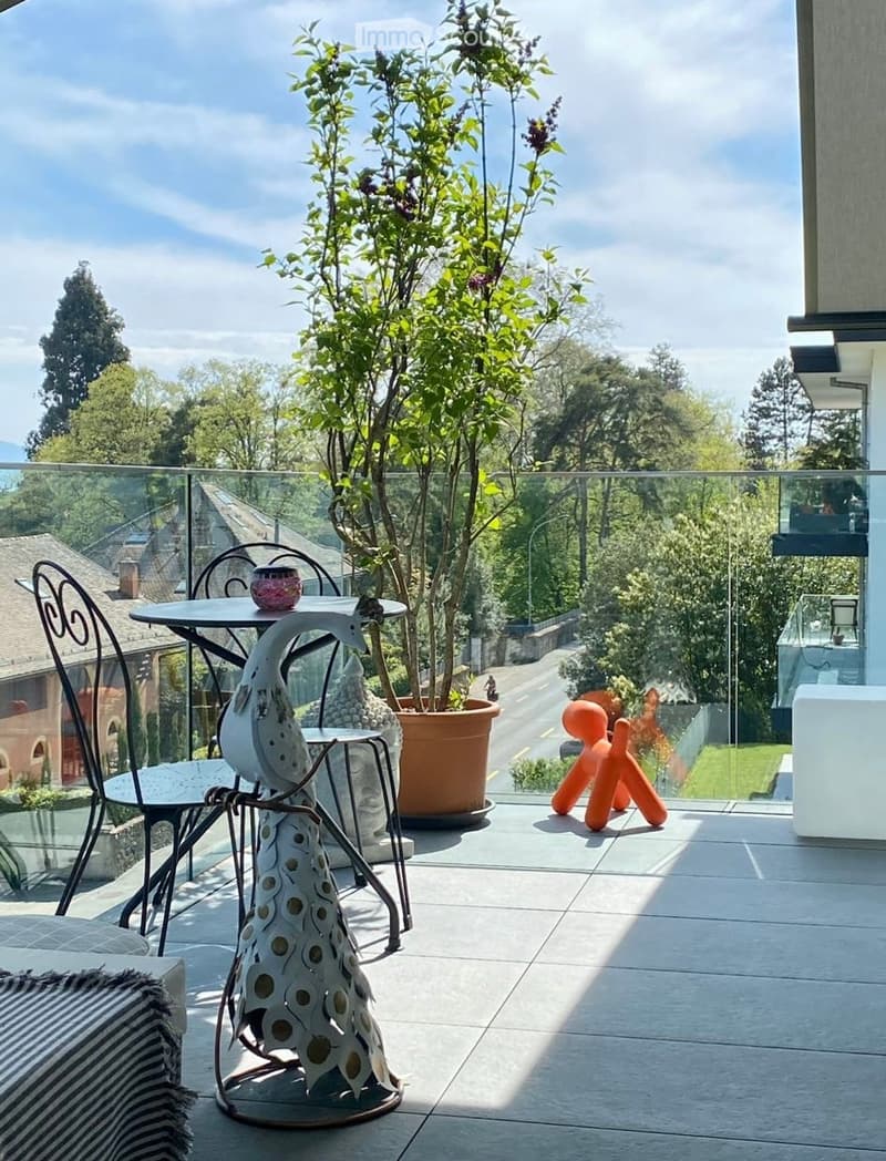 A modern balcony with a glass railing, featuring a small table, chairs, a decorative vase, a sculptural piece, and a potted plant. The view shows a tree-lined street and houses.