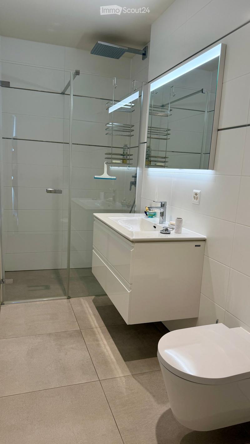 Modern bathroom with white tiles, a large mirror, a sink with a modern faucet, a toilet, and a glass-enclosed shower with a built-in shelf.
