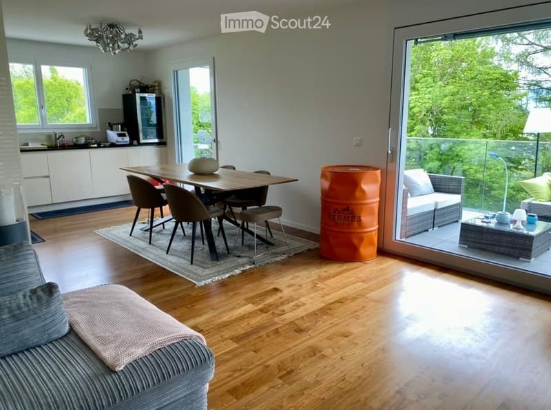 Open-plan living and dining area with wooden flooring, large windows, a sofa, coffee table, dining table with chairs, and a modern kitchen area with cabinets and a refrigerator.