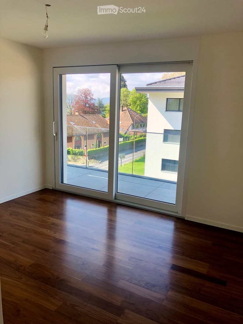 Empty room with wooden flooring and large sliding glass doors leading to a balcony or terrace, showing a view of the street and neighboring houses.