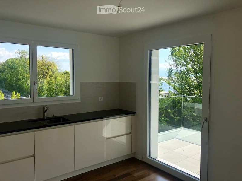 Modern kitchen with white cabinets, black countertop, and a sink. Large windows and a sliding glass door lead to a balcony with a view of greenery.