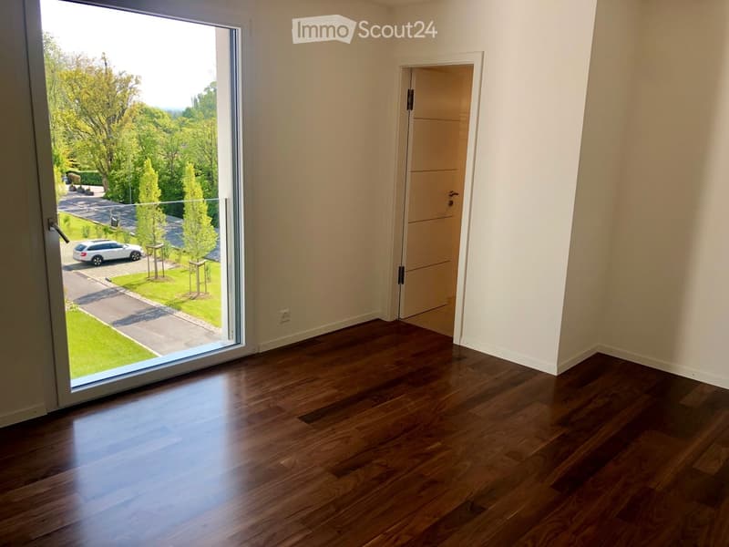 Empty room with wooden flooring, large sliding glass door leading to a green outdoor area, and a closed white door on the right side.