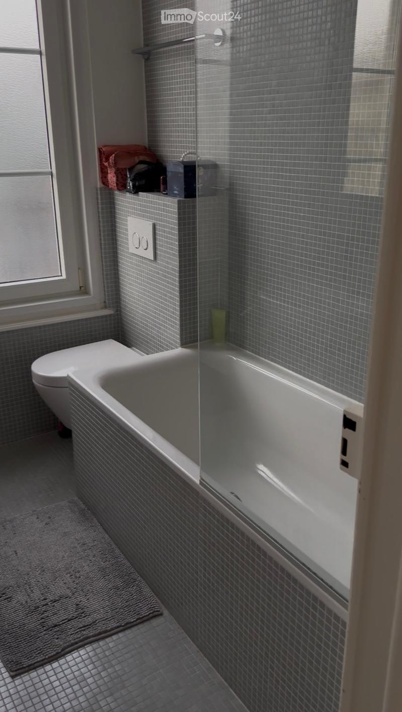 Modern bathroom with grey tiled walls and floor, featuring a bathtub with a glass shower enclosure, a toilet, and a towel on the shelf.