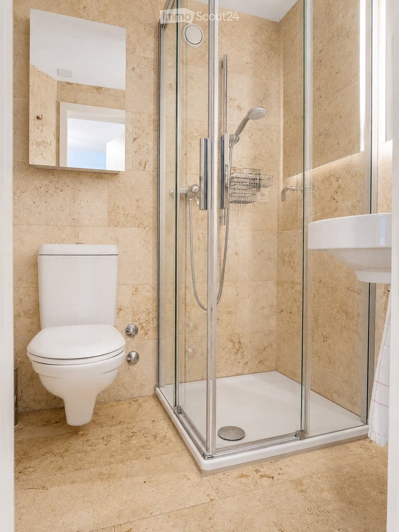 Modern bathroom with beige tiled walls and floor, featuring a white toilet, a glass-enclosed shower with a handheld showerhead, a white sink, and a mirrored cabinet above the sink.