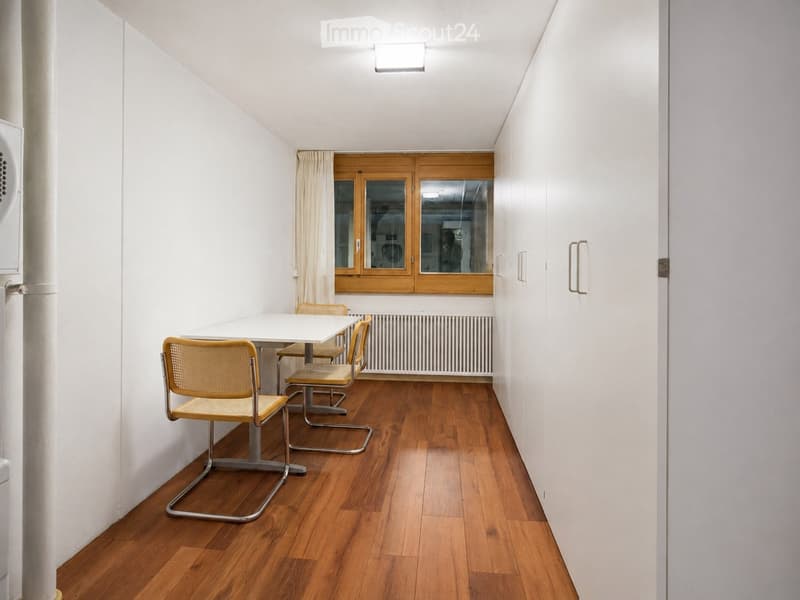 Bright, minimalist room with wooden flooring, white walls, and a small dining setup including a table and chairs. Features large windows with wooden frames and a radiator below them.