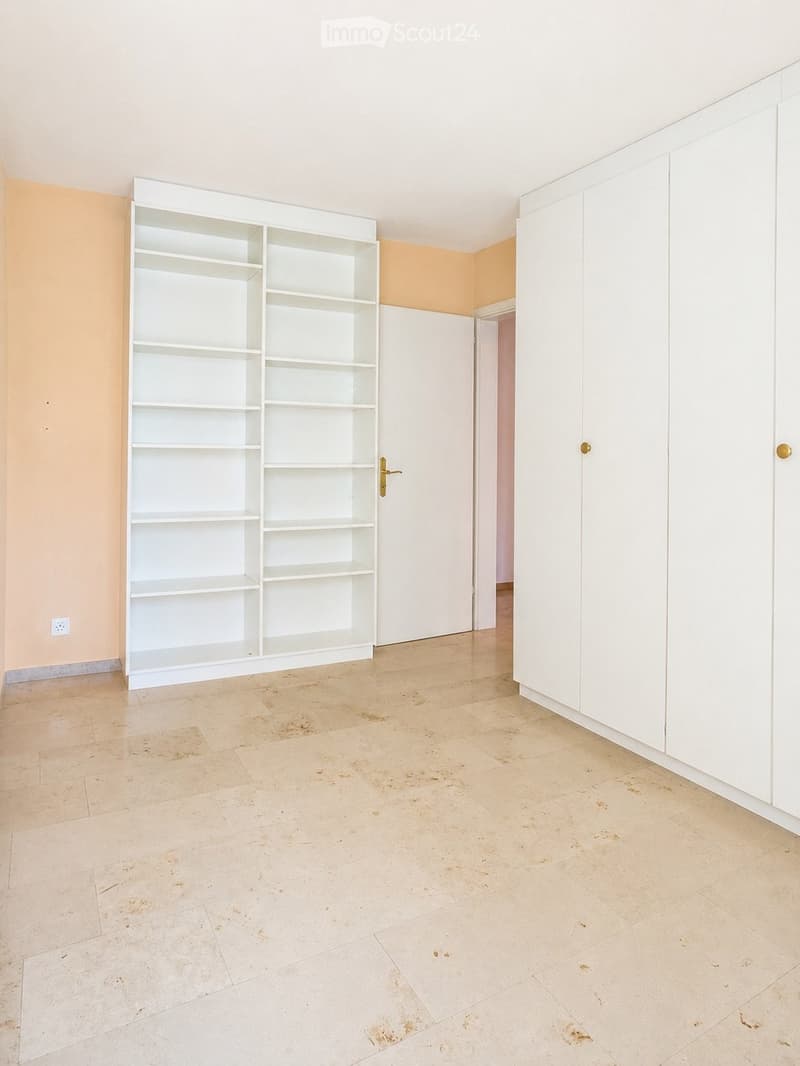 Empty room with beige tiled floor, white built-in shelving unit, and white cabinetry. The walls are painted in a light peach color, and there is a doorway leading to another room.