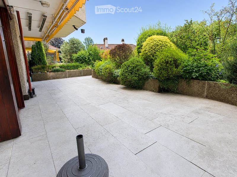 A well-maintained garden area with neatly trimmed bushes and trees, a tiled patio, and a small round table with a central pole.