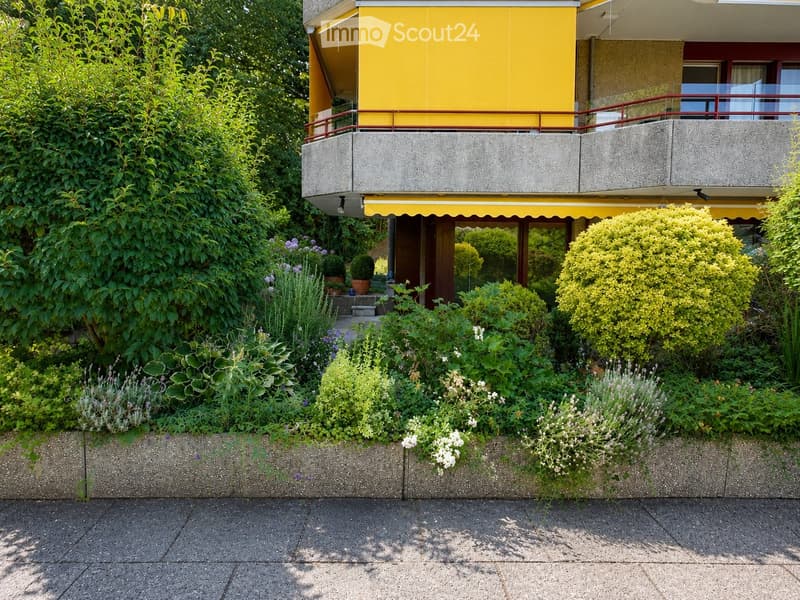 A well-maintained garden area with various plants, shrubs, and flowers, featuring a modern building with a yellow facade and balcony in the background.