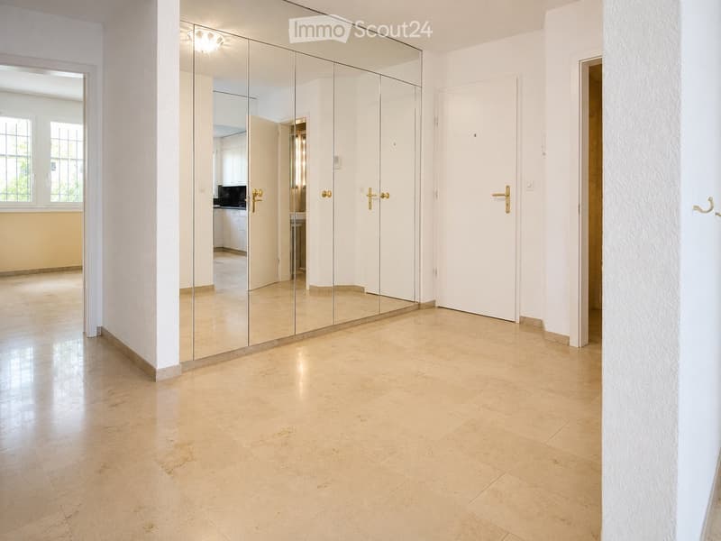 Bright hallway with beige tiled flooring, white walls, and large mirrored cabinet. Features multiple doors and a window on the left side.