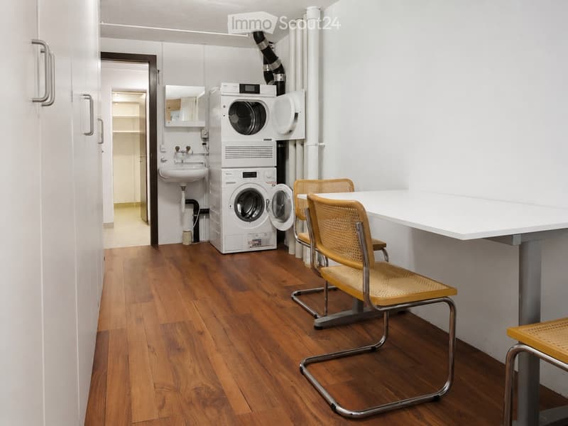 A small, well-lit room with wooden flooring, a washing machine and dryer unit, a sink, and a small seating area with chairs and a table.