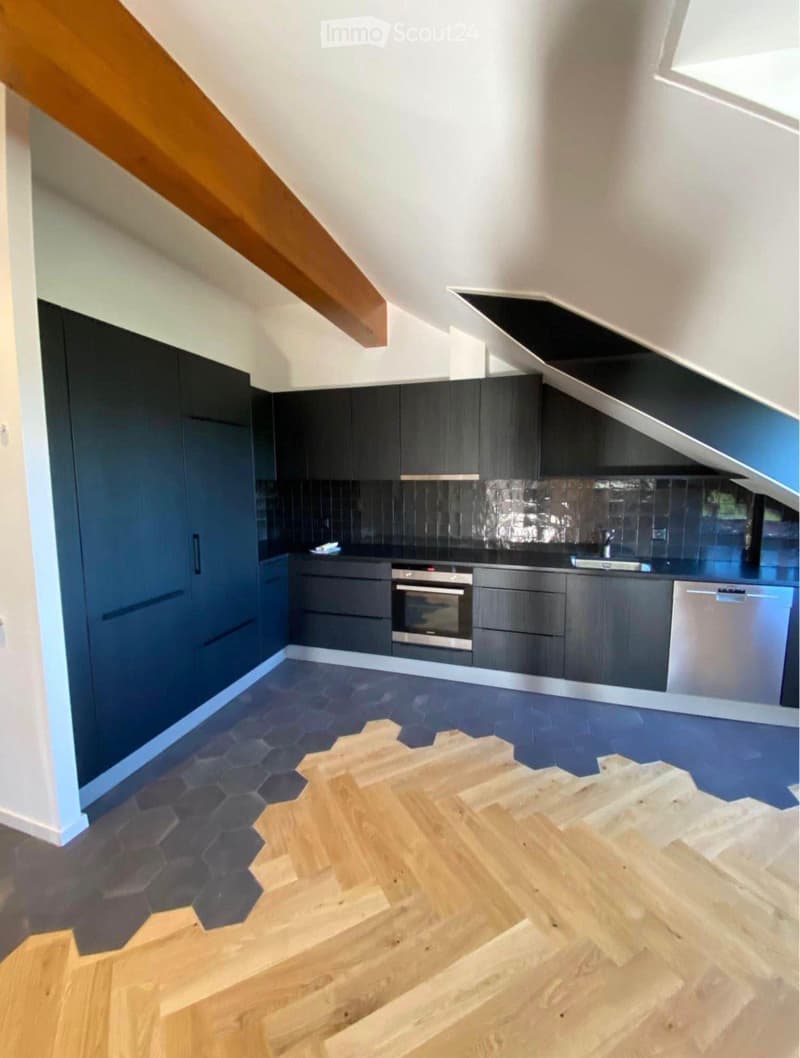 Modern kitchen with dark grey cabinets, stainless steel appliances, and a sleek countertop. The floor features a mix of hexagonal tiles and wooden planks, with a wooden staircase leading upwards.