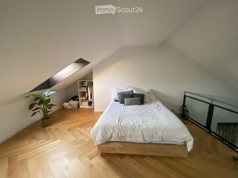 Cozy attic bedroom with wooden flooring, a large bed with white bedding and multiple pillows, a wardrobe, a potted plant, and a lantern on the floor near a glass railing.