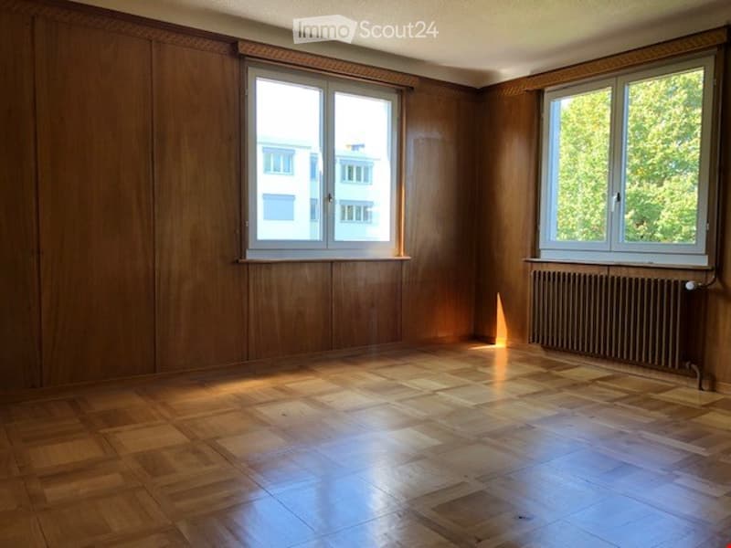 Empty room with wooden walls and floor, two windows, radiator, and natural light.
